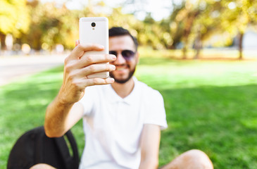 stylish hipster in a white shirt sits on the lawn in the Park and takes a selfie on your smartphone
