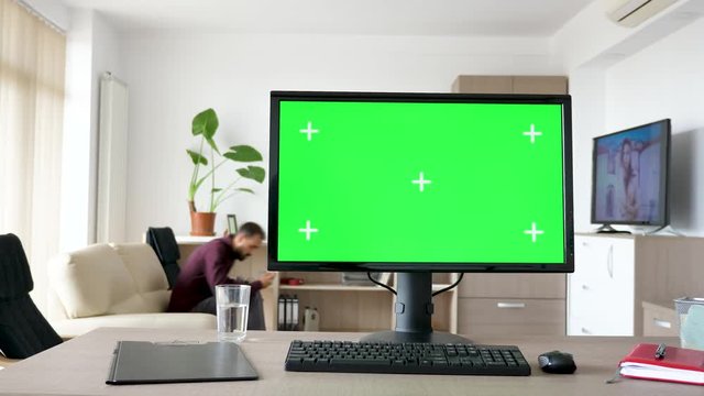 Modern personal computer with big green screen chroma mock up in the living room of comfy house. A man walks in background while the TV is on and sits on the sofa looking at the phone