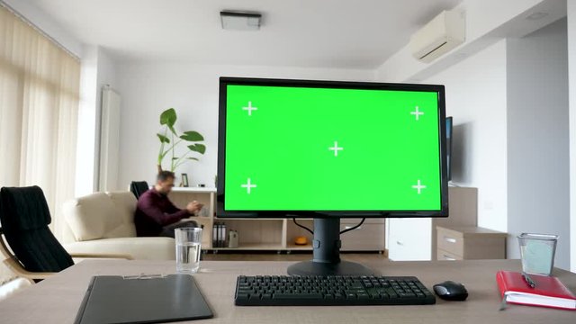 Dolly slider footage Personal PC computer with big green screen chroma mock up on the table in the living room. A guy is entering the room in background while the TV is on and sits on the sofa looking