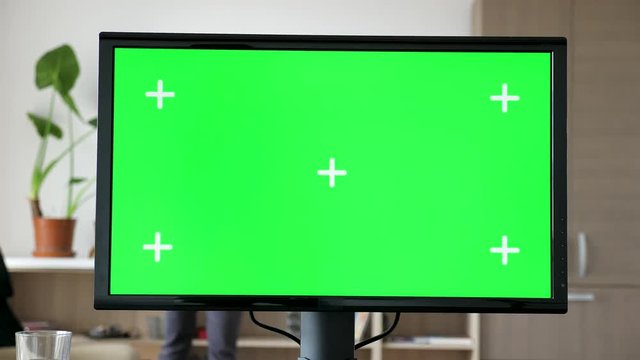 Modern Personal computer with big screen chroma mock up on the desk in living room. A man is entering the room in the background and sits on the couch while looking at his phone