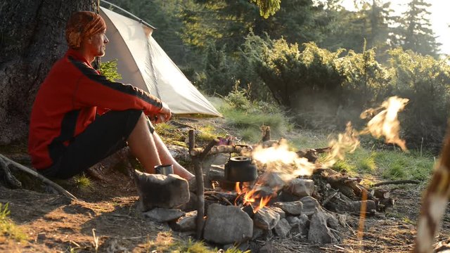 Happy adventurer drinks coffee and meditates next to the bonfire on a green mountain meadow next to tent - stunning morning during travel in the mountains.