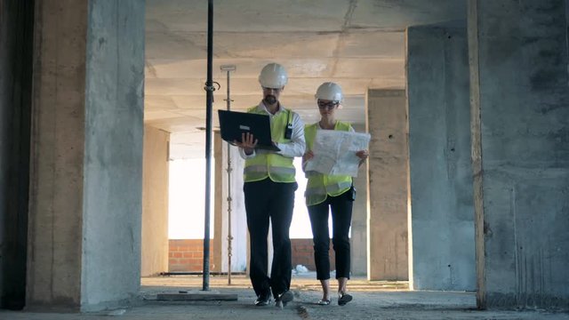 A female and a male construction workers, builders, constructors are walking along the construction site.