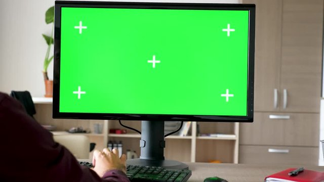 Side view of man sitting in front of computer with big green screen mock-up chroma typing on the keyboard. He is in the living room of his house. Dolly slider 4K footage with parallax effect