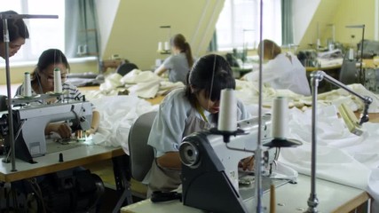 Group of multiethnic women using sewing machines to make clothes at factory, their boss controlling them