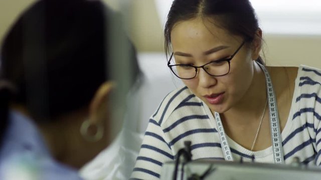 Tilt up medium shot of young Asian woman in glasses smiling and talking to colleague seen from her back when using sewing machine