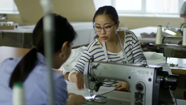 Medium shot of two Asian women talking to each other when using sewing machine to make clothes
