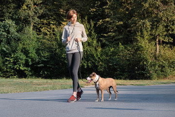 Woman trains her dog to run along during jogging. Young fit female and staffordshire terrier dog at...