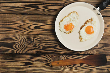 Two fried eggs in metal beige pan near dirty spatula on old wooden brown table. Top view with copy space