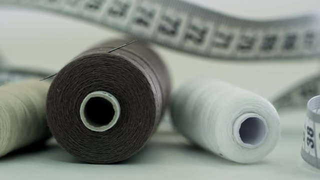Tracking shot of measuring tape and three spools of thread with needle stuck into one of them, lying on white background, close up