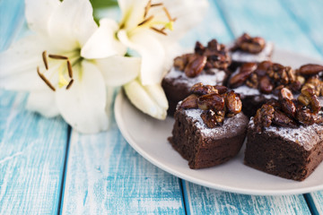 Pieces of chocolate cake with nuts on a plate next to the flowers of lilies.