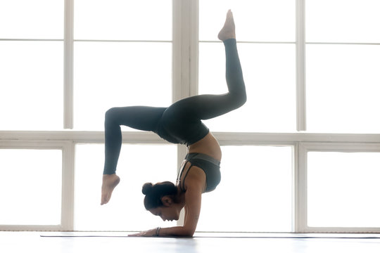 Young sporty attractive woman practicing yoga, doing Pincha Mayurasana exercise, handstand pose, working out, wearing sportswear, grey pants, top, indoor full length, at yoga studio