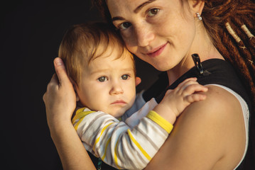 Portrait young happy mom with baby son in ergo backpack on dark wall on the background. Concept of modern parents