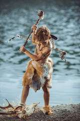 Angry caveman, manly boy with stone axe and bow hunting near river. Prehistoric tribal boy outdoors on nature. Young shaggy and dirty savage, warrior and hunter with weapon. Primitive ice age man in