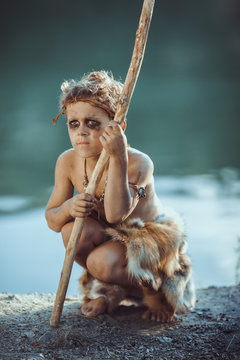 Sad caveman, manly boy with staff hunting. Prehistoric tribal boy outdoors on nature. Young shaggy and dirty savage, warrior and hunter. Primitive ice age man in animal skin sitting against river