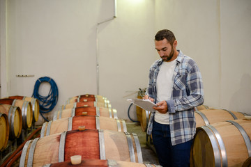Naklejka na ściany i meble handsome man winemaker in a maturing wine cellar winery during harvest season with oak barrels