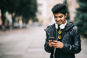 Happy indian guy walking and using a smart phone to listen music with headphones