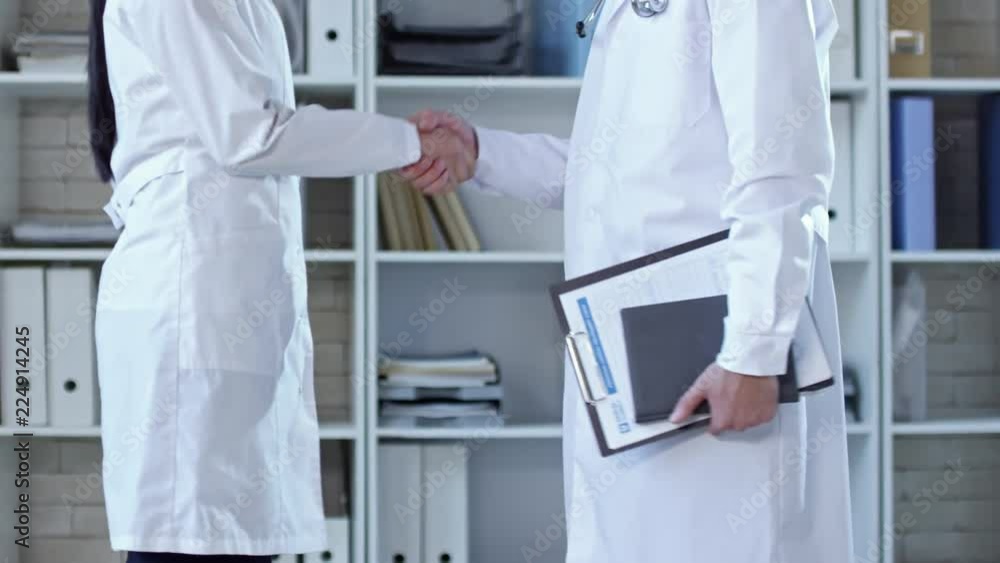 Poster side view of two unrecognizable doctors in lab coats approaching each other in office and shaking ha