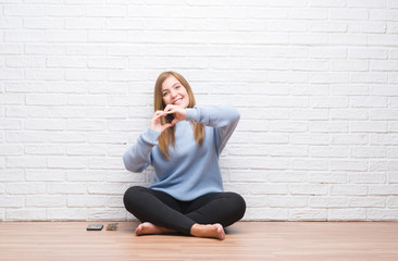Young adult woman sitting on the floor in autumn over white brick wall smiling in love showing heart symbol and shape with hands. Romantic concept.