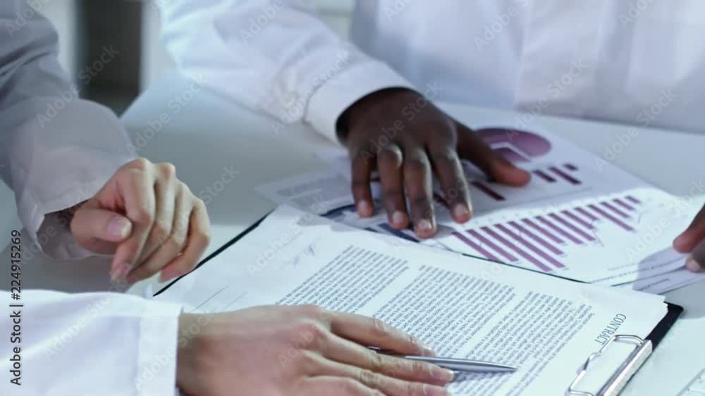 Poster Close up shot of two unrecognizable multi ethnic people in lab coats discussing documents and shaking hands when sitting at desk in office
