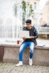 Obraz premium Handsome indian man with laptop while sitting near the fountain in the city center on a sunny summer day