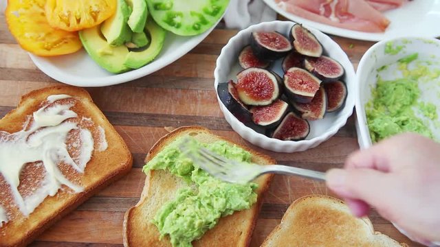 A woman adds mashed avocado, fresh figs and prosciutto to toast