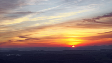 Colorful sunset in shades of yellow, red and orange with picturesque veil clouds in a hilly landscape, wide view