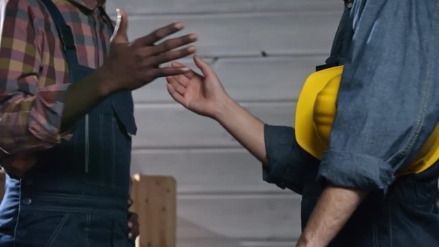 Tracking shot of two unrecognizable young men in overalls shaking hands after studying documents at work
