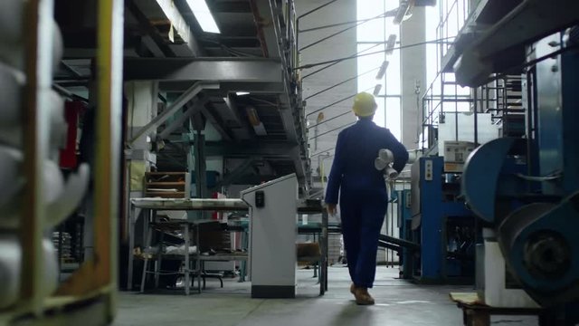 Tracking shot of factory worker in workwear and protective helmet holding rolled blueprints when walking through industrial facility, rear view