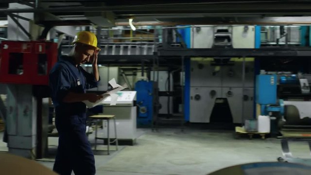 Follow shot of focused male factory worker walking through industrial building and trying to understand project papers, tracking right
