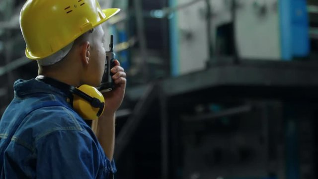 Tilt up shot of concentrated male factory worker in protective equipment operating machinery control panel and talking to colleagues via two way radio
