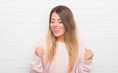 Young adult woman over white brick wall wearing winter outfit at home very happy and excited doing winner gesture with arms raised, smiling and screaming for success. Celebration concept.