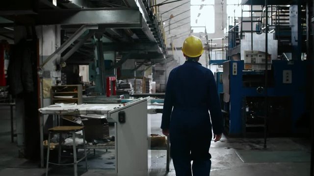Rear view follow shot of female engineer in yellow protective helmet walking though printing factory and taking sheets of paper