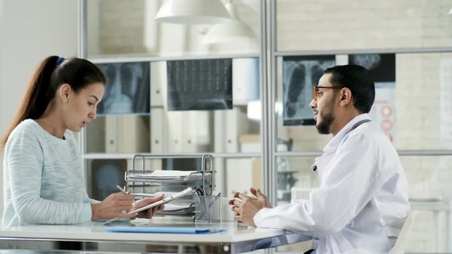 Side view of young Asian doctor in lab coat giving document attached to clipboard to female patient who is filling out form before health checkup