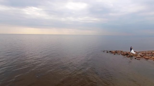 Bride and groom staying and hugging at the sandy beach rocks near sea at summer in wedding day. Aerial drone quadcopter shot at cloudy day.