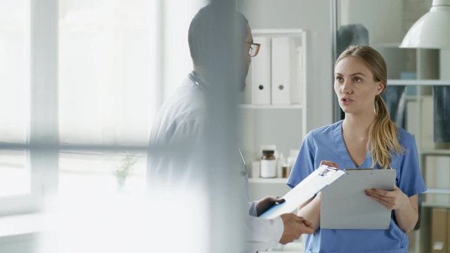 Tracking Medium Shot Of Two Young Doctors Or Scientists Of Different Ethnicities Holding Clipboards With Documents And Talking To Each Other At Work