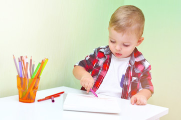 Cute little boy in a red checkered shirt smiles and draws a lilac marker in a white album
