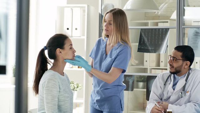 Tracking medium shot of two multiethnic doctors checking health of young female patient, writing down information and talking to her