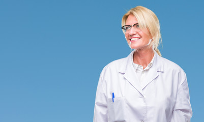 Middle age blonde therapist woman wearing white coat over isolated background looking away to side with smile on face, natural expression. Laughing confident.