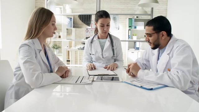 Zoom in shot of three multi ethnic young doctors in lab coats sitting at desk in medical clinic and discussing documents during meeting