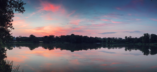 Fototapeta premium Calm evening lake scene with mirror like water reflections and purple pink sunset sky with moody green tree silhouettes. Braunschweig, Germany
