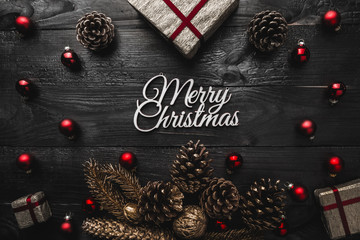 Upper, top, view from above, of evergreen pines, red toys, present and white Merry Christmas inscription on black wooden background, with space for text writing, greeting.