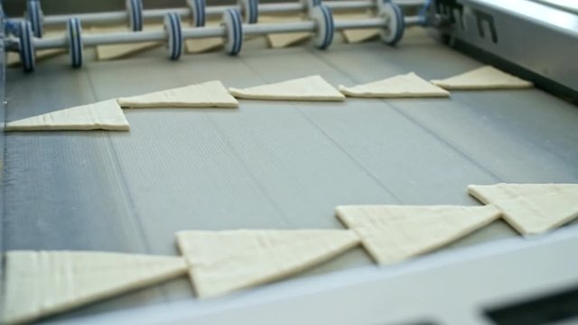 Close up shot of laminated dough for croissants being cut into triangular pieces on conveyor belt at factory