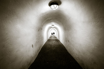 A long tunnel with white walls and arched ceiling. Two people are walking by the end of it.
