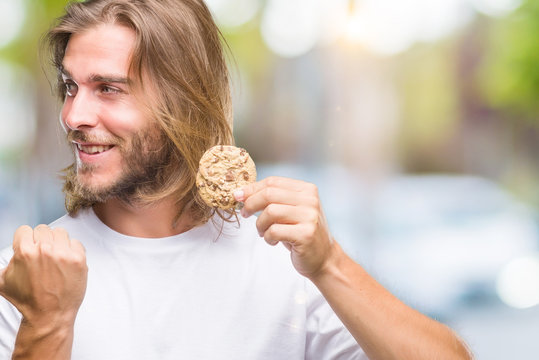 Young handsome man with long hair eating chocolate cooky over isolated background pointing and showing with thumb up to the side with happy face smiling