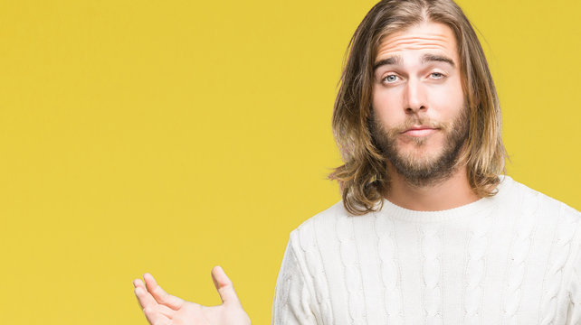 Young handsome man with long hair wearing winter sweater over isolated background clueless and confused expression with arms and hands raised. Doubt concept.