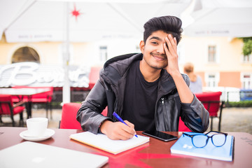 Young indian man freelancer of creative occupation writing down in copybook, noting goals, achievements and failures in time management check list during coffee break at cafe table