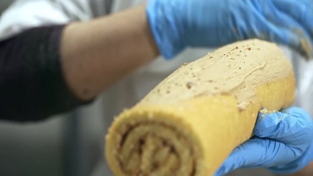 Close up shot of unrecognizable confectioner in blue gloves spreading delicious cream on swiss roll