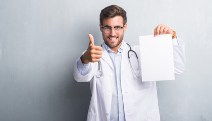 Handsome young doctor man over grey grunge wall holding blank sheet contract happy with big smile...