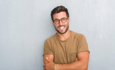 Handsome young man over grey grunge wall wearing glasses happy face smiling with crossed arms...