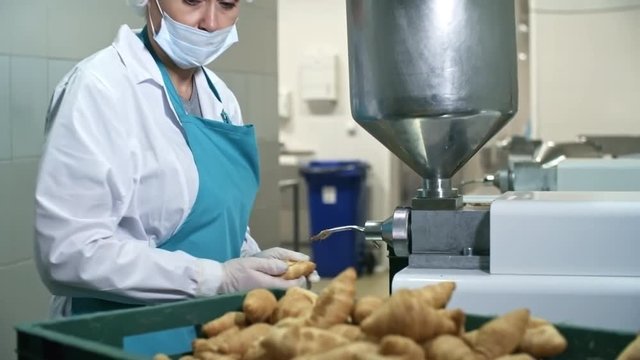 Tilt down shot of middle-aged aged female confectioner in protective clothing using filling machine to add cream to freshly baked croissants